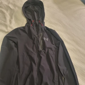 Svart jacka från Under Armour - Svart vindjacka från Under Armour med huva och röda detaljer vid snörningen. Jackan har dragkedja framtill, en bröstficka med dragkedja och diskret logga på bröstet. Inga byten. Se gärna mina andra annonser och skapa bundels.
