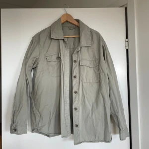 Beige overshirt från Urban Outfitters - Snygg beige overshirt från Urban Outfitters med klassisk krage, två bröstfickor med knapp och knäppning framtill. Perfekt att slänga över en t-shirt för en chill look. 