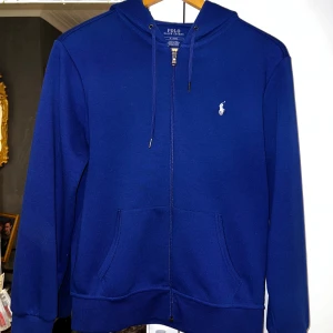 Blå hoodie från Polo Ralph Lauren - Snygg marinblå hoodie från Polo Ralph Lauren med dragkedja och klassisk vit logga på bröstet. Tröjan har huva med snörning, två fickor framtill och långa ärmar. Perfekt för en chill och stilren look.