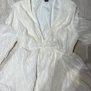 Vit omlottklänning med spetsdetaljer - Supersöt vit blus med omlott och v-ringad front. Blusen har långa ärmar i transparent spets med ett diskret mönster och markerad midja. Perfekt för dig som gillar romantisk och feminin stil. Materialet känns lätt och luftigt.