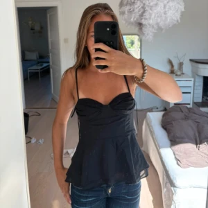 Svart korsett-topp med volang - Snygg svart korsett-topp med tunna axelband som knyts och en volang nedtill. Toppen har en figurnära passform och markerad byst med lätt rynkade detaljer. 