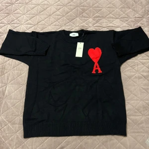 Svart stickad tröja från Ami - Svart stickad tröja från Ami Alexandre Mattiussi med rund hals och ribbade muddar. På bröstet finns en stor röd hjärtapplikation med bokstaven A. Perfekt för dig som gillar stilrena plagg med en twist.