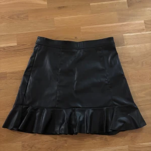 Svart kort kjol med volang i skinn - Snygg svart kort kjol i skinnimitation med bred volang längst ner. Kjolen har en stilren design och en dragkedja bak för enkel på- och avtagning. Perfekt för dig som gillar edgy och trendiga plagg med lite attityd.