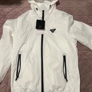 Vit vindjacka från Vogue Style - Snygg vit vindjacka med svarta detaljer från Vogue Style/Fashion. Jackan har huva, dragkedja framtill och två fickor med svarta dragkedjor. På bröstet sitter en svartvit trekantig logga med texten 'Fashion'. Perfekt för dig som gillar clean och sportig stil.