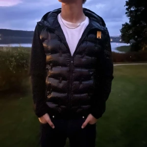 Svart Cardigan från Parajumpers - Säljer en svart cardigan från Parajumpers med glansig nylon på fram- och baksidan samt ärmar i mjuk fleece. Jackan har dragkedja framtill, huva och en gul detalj vid axeln. Perfekt för dig som vill ha en stilren look. Använd fåtal gånger på grund av att den inte passa min stil. Kan sänka priset vid snabba affärer