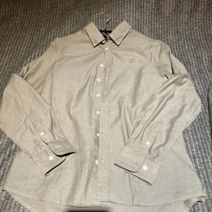 Beige grön bomullsskjorta från Gant - Klassisk beige skjorta från Gant i bomull med broderad logga på bröstet. Skjortan har lång ärm, knappar framtill och på manschetterna samt en snygg krage. Perfekt för en clean och stilren look.
