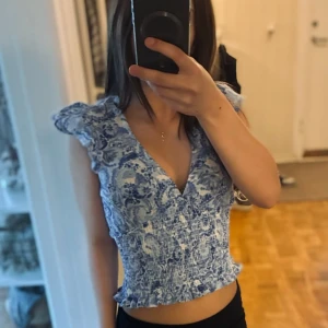 Blommig blåvit blus från Gina Tricot - Supersöt blommig blus från Gina Tricot i blått och vit!❤️Modellen är croppad med smockad midja, volangdetaljer vid axlar och nederkant samt v-ringning. Perfekt för dig som gillar blå vibes och vill sticka ut lite extra.❤️