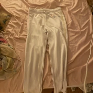 Vita mjukisbyxor från Champion L - Vita mjukisbyxor från Champion med elastisk midja och dragsko. Byxorna har färgade ränder längs sidorna, en gul och en rosa, samt broderad Champion-logga i flera färger vid fickan. Tillverkade i mjuk bomull, perfekta för chill dagar. Det finns ingen ficka och har två små hål. Ett på knät och ett vid midjan.