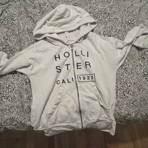 Vit hoodie med dragkedja från Hollister - Vit hoodie från Hollister med svart tryck framtill och texten 'Hollister Cali 1922'. Tröjan har huva med snörning, dragkedja och två fickor framtill. Perfekt för chill dagar och enkel att matcha med jeans eller leggings.