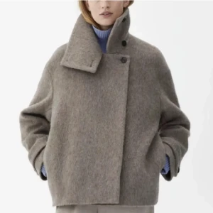 Beige ulljacka med hög krage - Säljer en snygg beige jacka i ull med oversized passform och hög krage som kan knäppas upp. Jackan har en asymmetrisk knäppning framtill och långa ärmar. Perfekt för dig som gillar stilrena och trendiga plagg.