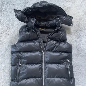 Svart dunväst från Leon Marcel - Snygg svart dunväst från Leon Marcel med avtagbar huva och två dragkedjefickor framtill. Västen har quiltad design och blanka paneler som ger en modern look. Perfekt att slänga över en hoodie för extra värme under kyliga dagar. Använd en gång och endast provad.