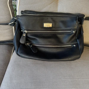 Svart axelväska från Feel i skinn - Snygg svart axelväska från Feel i skinnimitation med flera dragkedjefack både utvändigt och invändigt. Väskan har justerbar axelrem och ett mönstrat innerfoder. Perfekt för dig som vill ha ordning på dina grejer och gillar stilren design.