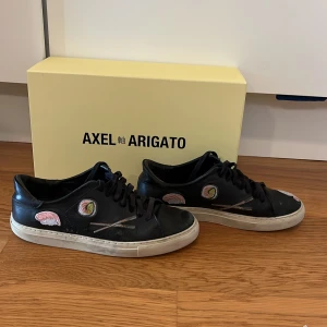 Svarta Axel Arigato sneakers med sushi-broderi - Unika svarta sneakers från Axel Arigato med broderade sushi-motiv och vita sulor. Box och påse ingår. Skriv vid funderingar eller om du har en fråga!