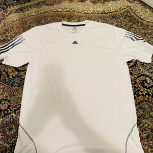 Vit Adidas t-shirt i polyester - Säljer en vit Adidas t-shirt i polyester med svarta detaljer på axlarna och logga på bröstet. T-shirten har korta ärmar och sportig design, perfekt för träning eller chill. Snygga sömmar längs sidorna för extra stilpoäng.