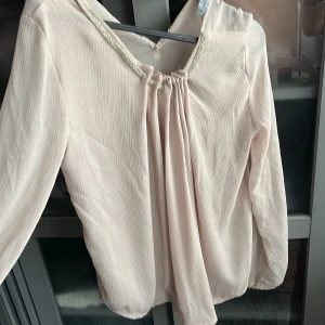 Ljusrosa blus från Rinascimento - Supersöt ljusrosa blus från Rinascimento med plisserat tyg och långa ärmar. Blusen har en glittrig detalj längs halsringningen och ett lätt, luftigt material som ger en flowy känsla. Perfekt för dig som gillar eleganta och feminina plagg.