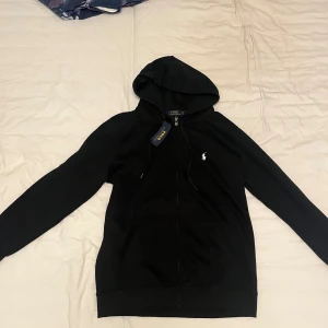 Svart hoodie  - Svart hoodie med klassisk vit broderad logga på bröstet. Tröjan har huva med snörning, dragkedja framtill och känguruficka. Tillverkad i mjuk bomull, perfekt för en chill och stilren look.