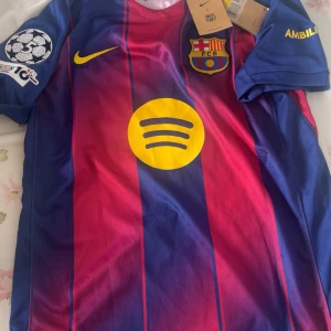  Barcelona Lamine Yamal matchtröja - Säljer en officiell FC Barcelona matchtröja från Nike med Lamine Yamal och nummer 10 på ryggen. Tröjan är randig i blått och rött med gula detaljer, klubbmärke och Champions League-patch på ärmen. Materialet är lätt och andas, perfekt för fotboll eller supporterliv.