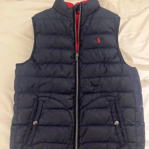 Vändbar dunväst från Ralph Lauren - Snygg vändbar dunväst från Ralph Lauren med ena sidan marinblå och andra sidan röd. Västen har dragkedja framtill, två fickor med knapp och klassisk logga på bröstet. Perfekt för lager-på-lager och funkar till många olika outfits. Storleken är 14-16 år d.v.s barn L men sitter som XS-S