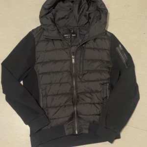 Svart pufferjacka Supply & Demand L - Svart pufferjacka från Supply & Demand med huva och dragkedja framtill. Jackan har quiltad framsida och rygg upptill, släta ärmar och en ficka med dragkedja på ena ärmen. Perfekt för dig som gillar streetwear och vill ha en stilren look. Pris kan diskuteras! 