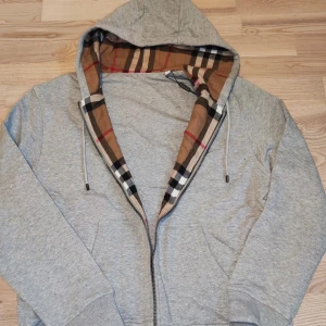 Grå Burberry hoddie  - Grå hoodien från Burberry med klassiskt rutigt foder i huvan och broderad logga på bröstet. Jackan har dragkedja, snörning i huvan och två fickor framtill. Perfekt för dig som gillar stilren design med en touch av lyx.