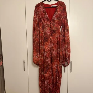 Röd blommig långklänning Adore - Säljer en maxad röd långklänning från Adore med blommigt mönster och djup v-ringning. Klänningen har långa puffärmar med knappar vid ärmslut och en snygg knappdetalj framtill. Perfekt för dig som vill sticka ut med färg och mönster.