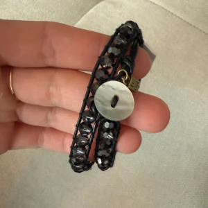 Armband  - Snyggt armband från med två varv av mörkgrå pärlor på ett svart läderband. Armbandet knäpps med en vit pärlemorknapp. En liten berlock med texten "IC ON" hänger vid knäppningen.