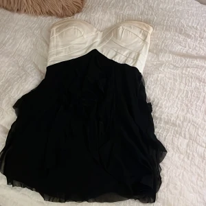 Svart och vit axelbandslös kortklänning - Säljer en gammal mini klänning med svarta volanger. Där bak är det en zip som man knäpper den med. Den har en elegant, festlig vibe. 