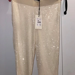 Glittriga paljettbyxor i beige - Snygga beige byxor från PS Bling med massor av paljetter som ger ett riktigt glittrigt intryck. Byxorna har elastisk midja och är täckta av små, skimrande paljetter för en extra festlig look. Perfekta för dig som vill sticka ut.