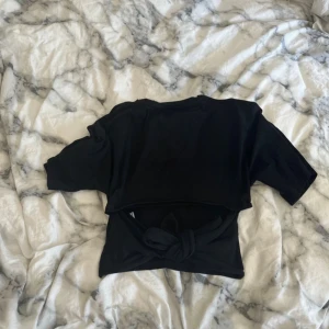 Svart croppad topp med knyt - Svart croppad topp med korta ärmar och breda knytband i midjan som kan knytas framtill eller baktill för en cool look. T-shirttyg i bomull, rund halsringning och enkel stil som passar perfekt till jeans eller kjol.