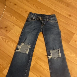 Blå slitna bootcut jeans - Säljer ett par blå jeans med bootcut passform och stora slitningar över knäna. Jeansen har låg midja, klassiska fem fickor och är gjorda i denim. Perfekta för en avslappnad och trendig look.