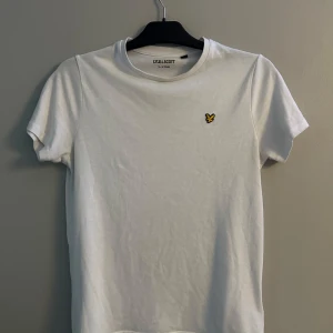 Vit t-shirt från Lyle & Scott - Vit t-shirt från Lyle & Scott med klassisk passform och korta ärmar. T-shirten har rund halsringning och en gul  logga på bröstet. Tillverkad i mjuk bomull, perfekt för en clean och stilren look. Ordinarie pris 300kr