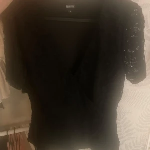 Svart spetsblus från BubbleRoom - Svart blus från BubbleRoom med omlott-skärning och korta ärmar i genombruten spets. Snygg v-ringning och figurnära passform, perfekt att styla med jeans eller kjol. Spetsdetaljerna ger en extra touch till outfiten.