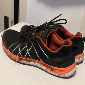 Svarta och orange sneakers från Gesto - Snygga svarta sneakers från Gesto med orange detaljer och meshfoder. Skorna har snörning, vit och orange sula samt reflekterande mönster på sidorna. Perfekta för dig som gillar sportig stil och vill sticka ut med färg. Lätta och bekväma för träning eller vardag.