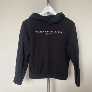Svart hoodie från Tommy Hilfiger - Svart hoodie från Tommy Hilfiger med broderad logga och text på bröstet. Klassisk huva och långa ärmar, ribbade muddar och nederkant. Perfekt för en chill och stilren look.