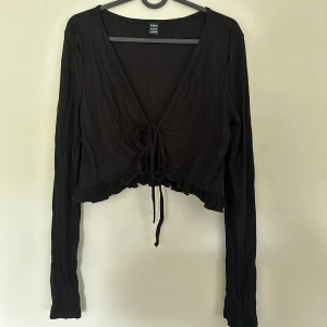 Svart croppad bolerotopp med snörning - Svart croppad bolerotopp från SHEIN med långa ärmar, v-ringning och snörning framtill. Toppen har ribbad struktur och volangkant nedtill för en extra söt touch. Storlek L men passar M/S