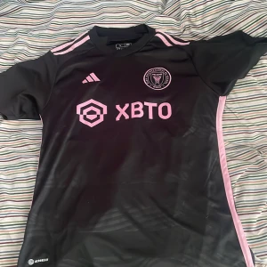 Inter Miami svart fotbollströja Adidas - Svart Inter Miami fotbollströja från Adidas i slim fit med rosa detaljer och klubbmärke på bröstet. Tröjan har korta ärmar, rosa ränder på axlarna och tryckt sponsorlogga framtill. Tillverkad i lätt och ventilerande material för max komfort.
