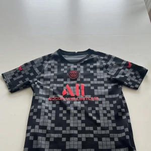 PSG x Nike svart-rutig fotbollströja - Snygg PSG fotbollströja från Nike i svart med grått rutmönster och röda detaljer. Tröjan har korta ärmar, rund hals och är gjord i lätt polyester som andas. Paris Saint-Germain-logga på bröstet och tryck både fram och bak. Perfekt för dig som älskar fotboll och streetstyle. Storlek barn XL