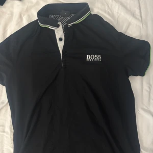 Svart Hugo Boss t-shirt med krage - Svart t-shirt från Hugo Boss med vit knappslå och krage med gröna och vita detaljer. Kortärmad modell med BOSS-logga broderad på bröstet. Snyggt mönstrad insida vid halsen och klassisk passform. Perfekt för dig som gillar stilrena märkesplagg.
