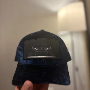 Blå BMW M5 E60 truckerkeps - Snygg marinblå truckerkeps med sammetsliknande front och mesh på sidorna och bak. Framtill finns en patch med motiv av BMW M5 E60. Justerbar snapback för perfekt passform. Perfekt för dig som gillar bilar och vill sticka ut med en unik keps. Jag säljer denna pågrund av fel passform. Den är helt oanvänd och helt ny kartong följs med, Original pris 400kr.