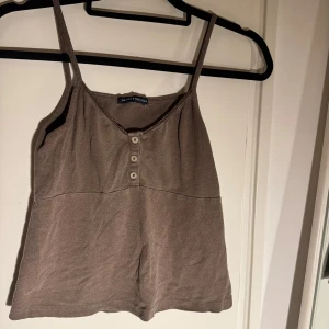 Brun linnetopp från Brandy Melville - Supersöt brun linnetopp från Brandy Melville med smala axelband och fyra knappar framtill. Toppen har en lätt v-ringning och diskret spetskant längs urringningen. Perfekt för en chill och avslappnad stil.