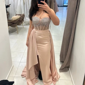 Champagnefärgad långklänning med pärlor - Säljer en elegant långklänning i champagnefärg med glittrig pärlbeströdd överdel och smala axelband. Kjolen är i satin med draperad detalj och hög slits framtill. Perfekt för bal eller bröllop. Överdelen har en snygg v-ringning och markerad midja. Jag har tagit bort banden vid brösten och gjort dom så som på andra bilden! Använd endast en gång.