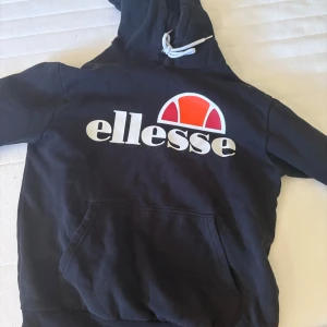 Svart Ellesse hoodie med logga - Svart hoodie från Ellesse med stor vit och röd logga framtill. Tröjan har huva med snörning och en klassisk känguruficka. Perfekt för en avslappnad och sportig stil.