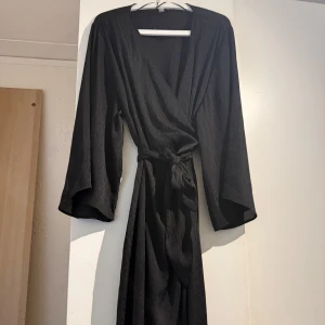 Svart omlottklänning från H&M - Svart långklänning från H&M i omlottmodell med knyt i midjan. Klänningen har vida långärmar och ett lätt, glansigt tyg som ger ett elegant intryck. Perfekt för dig som gillar stilrena och tidlösa plagg med snygg passform.