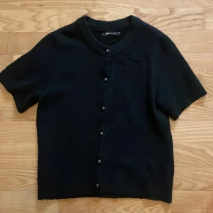 Svart kofta med kort ärm från Gina Tricot - Kortärmad svart kofta från Gina Tricot med rund halsringning och knappar framtill. Tröjan är stickad och har en klassisk och stilren design med mjukt material.