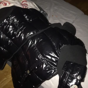 Svart dunväst från Moncler - Säljer en svart, glansig dunväst från Moncler med ribbad krage och muddar. Västen har quiltad design, dragkedja framtill och tryckknappar på fickorna. Perfekt att slänga över en hoodie för extra värme och stil. Passar m och s