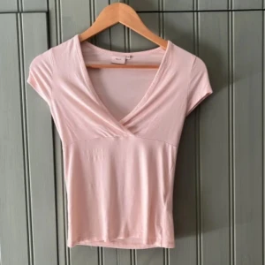 Ljusrosa V-ringad topp från Nelly🩷 - Supersöt ljusrosa topp från Nelly med omlottdesign och v-ringning. T-shirtrmar och figurnära passform gör den perfekt till jeans eller kjol. Mjuk och stretchig i bomullsmix, enkel att styla för en fräsch look.