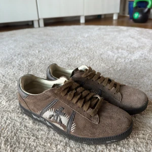 Premiata skor - Snygga bruna sneakers från Premiata i mjuk mocka med broderad logga på sidan och svarta detaljer. Skorna har snörning, svart platt sula och rund tå. Perfekta för dig som gillar en stilren men unik look. Storlek 40 men passar större.