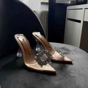 Glittriga pumps med  klack - Snygga pumps med spetsig tå i beige och transparent material. Skorna har en unik genomskinlig klack och är dekorerade med stora glittriga stenar framtill. Använda ett par gånger men i gott skick 