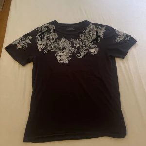 Svart t-shirt med barockmönster från Zara - Svart t-shirt från Zara med grymt snyggt barockinspirerat mönster i vitt och grått över axlar och bröst. Klassisk rund halsringning och korta ärmar. Materialet är mjuk bomull som känns skönt mot huden. Perfekt för dig som vill sticka ut lite extra.