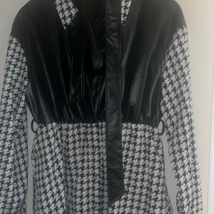 Svartvit skjorta med skinnimitation - Unik skjorta med svart skinnimitation upptill och klassiskt svartvitt hundtandsmönster nedtill och på ärmarna. Långärmad modell med krage och knytband framtill. Perfekt för dig som vill sticka ut med en cool och trendig look.